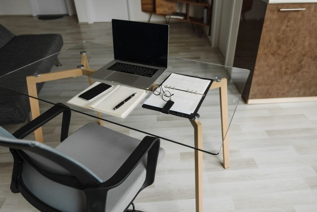Bureau privé lumineux avec plan de travail en verre, ordinateur portable, carnet et lunettes posés dessus